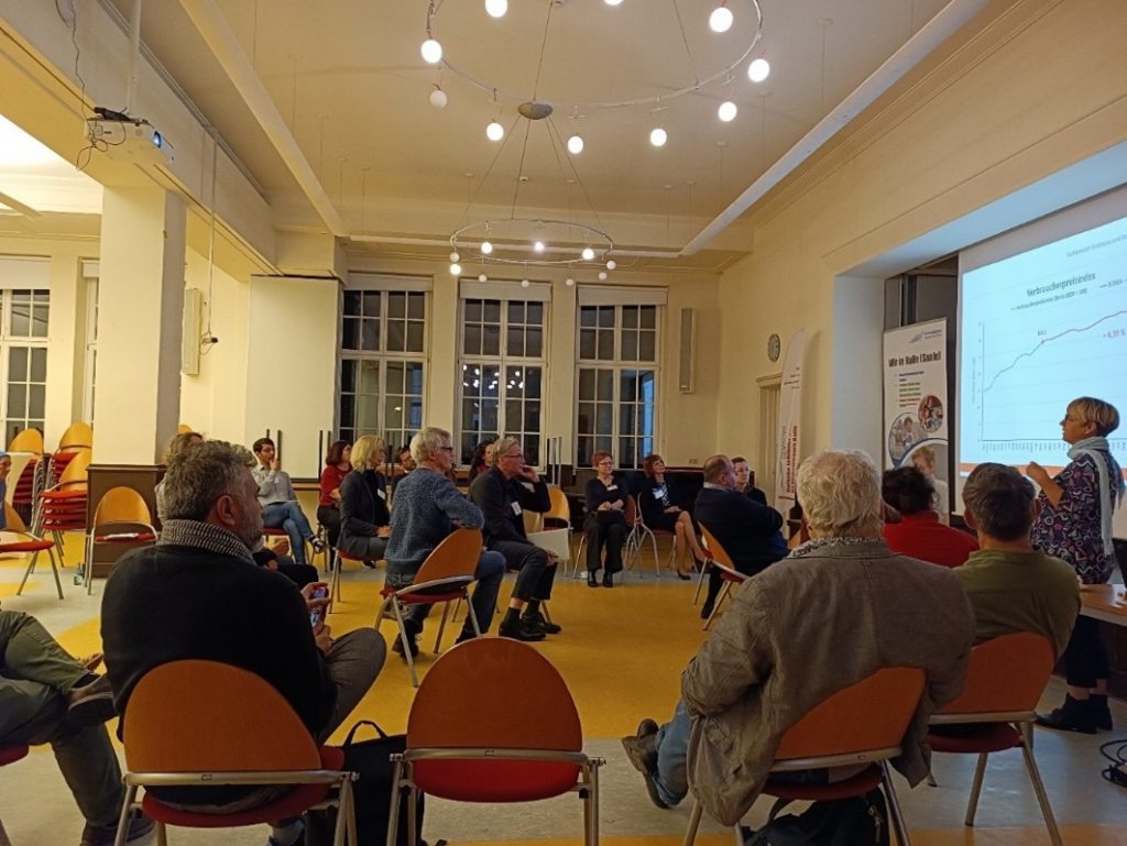 Zu sehen sind eine Runde von menschen die auf dem Stühlen sitzen und einer Präsnetation zuhören.