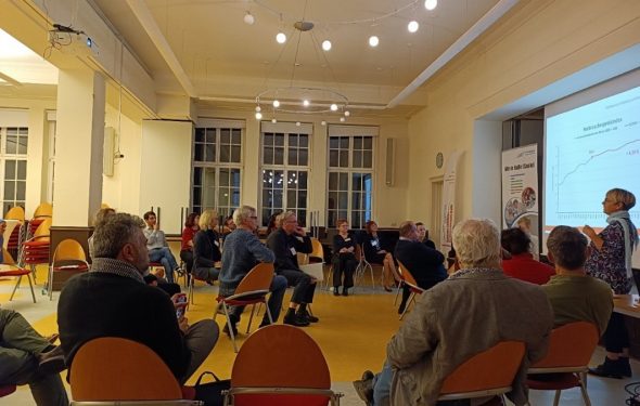 Zu sehen sind eine Runde von menschen die auf dem Stühlen sitzen und einer Präsnetation zuhören.