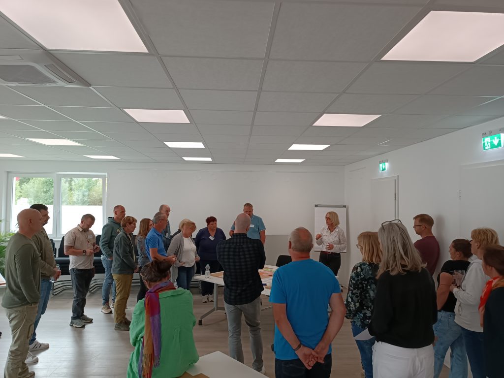 Zu sehen sind viele Personen die im Kreis stehen und durch eine moderierende Person bei einer Workshop Veranstaltung unterstützt werden.