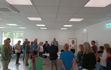 Zu sehen sind viele Personen die im Kreis stehen und durch eine moderierende Person bei einer Workshop Veranstaltung unterstützt werden.