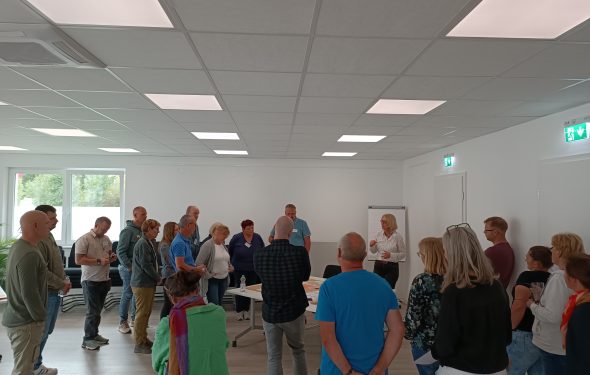 Zu sehen sind viele Personen die im Kreis stehen und durch eine moderierende Person bei einer Workshop Veranstaltung unterstützt werden.